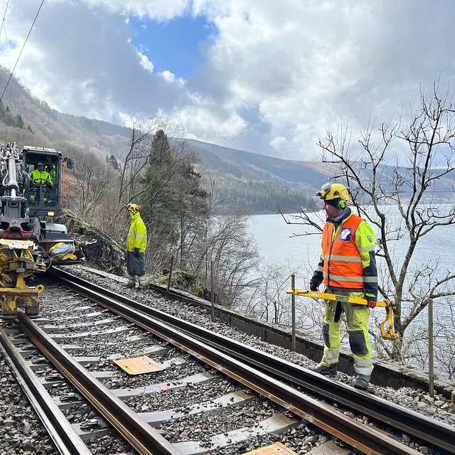 Jernbanearbeid med maskin langs spor ved innsjø og fjell