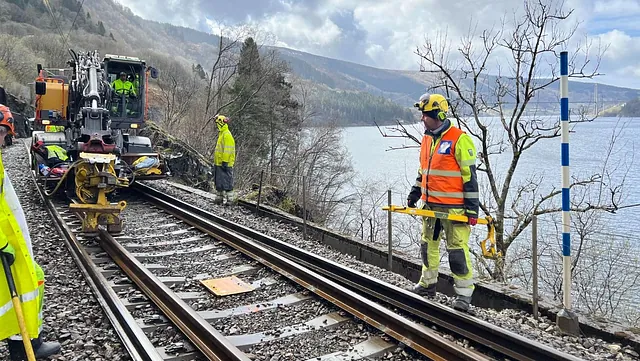 Jernbanearbeid med maskin langs spor ved innsjø og fjell