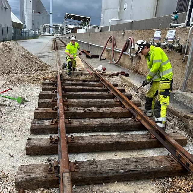 To arbeidere monterer jernbaneskinner i industriområde