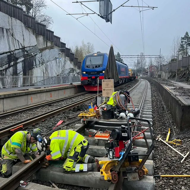 Arbeidere utfører vedlikehold på jernbanespor med tog i bakgrunnen